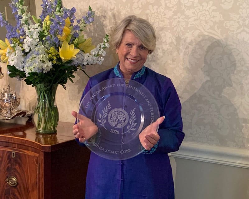 Blonde short haired woman in a royal purple button up jacket holds a glass plate that reads: The NSCDA Award for Generosity and Purpose 2020, Virginia Stuart Cobb