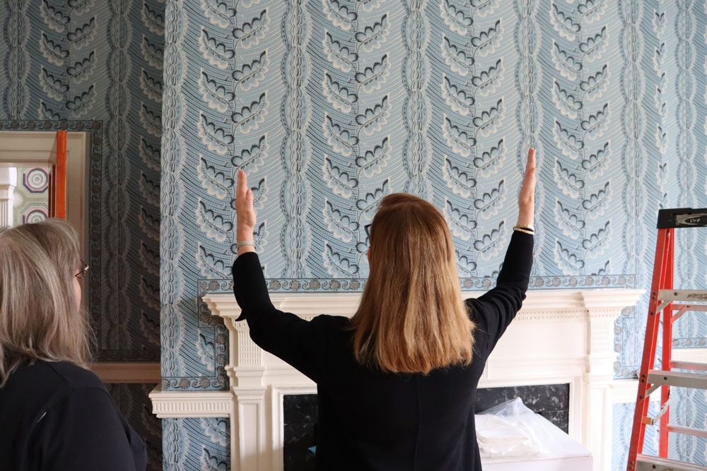 Woman with shoulder length red hair gestures to a wall covered in light blue patterned wallpaper