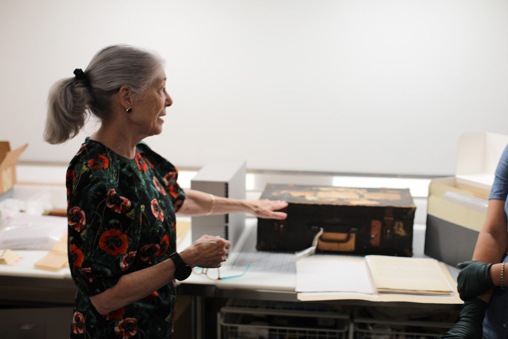 Woman with silver hair in a floral dress indicates an old looking suitcase on a table surrounded by files