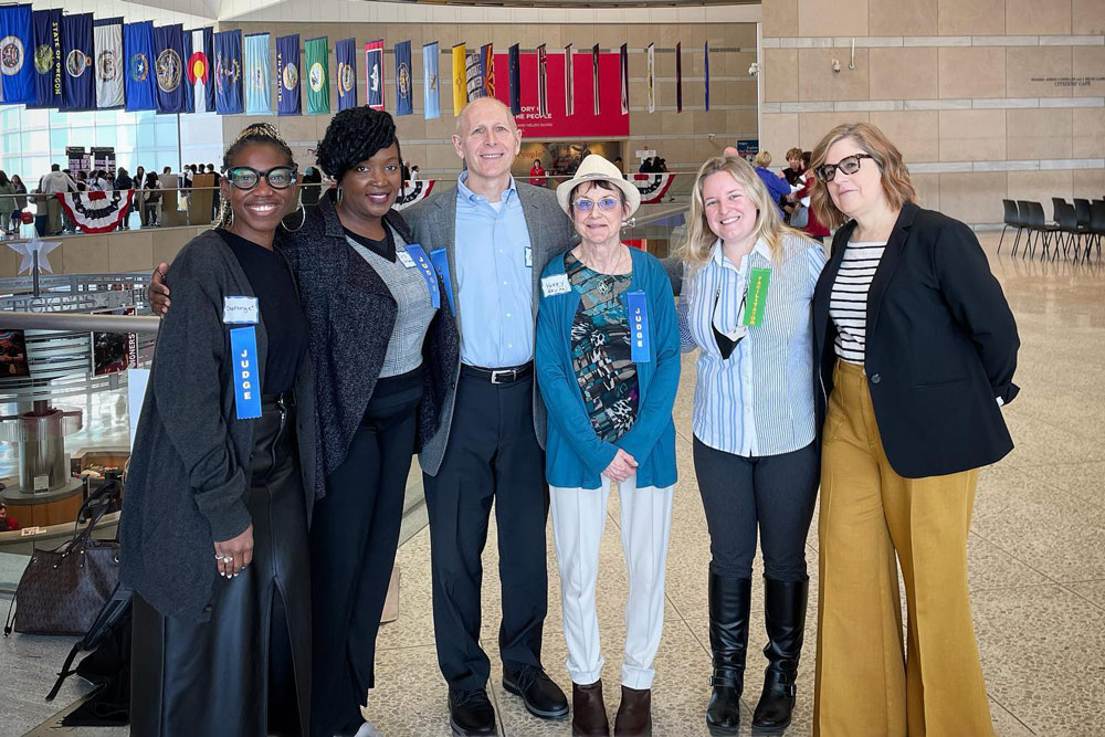 Competition judges stand in a line and smile at the camera, five women and one man