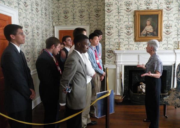 Group of teenagers stand behind a golden rope and listen to a silver-haired woman talk