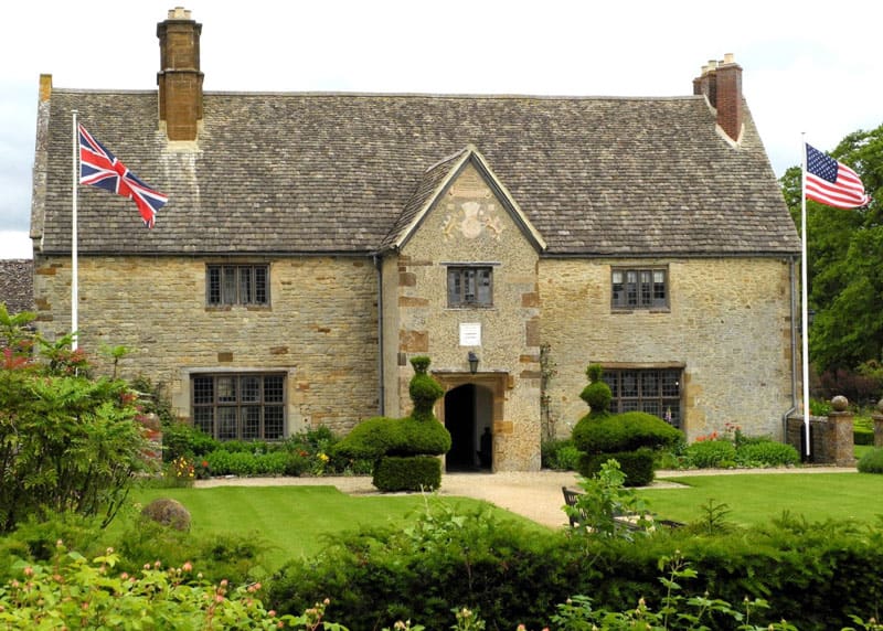 Exterior shot of Sulgrave Manor, a stone house with manicured shrubs and lawn and both a UK and US flag flying in the front