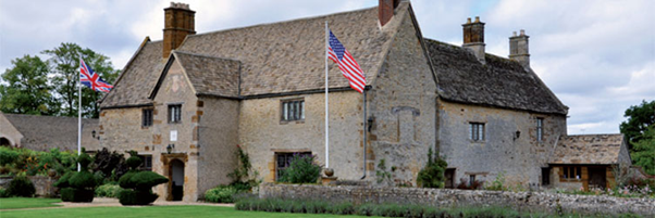 Exterior shot of Sulgrave Manor, a stone house with manicured shrubs and lawn and both a UK and US flag flying in the front