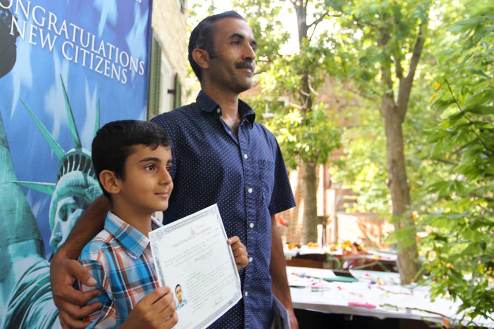 Older man with short sleeved navy button down and mustache puts arm around shoulders of young boy holding a certificate