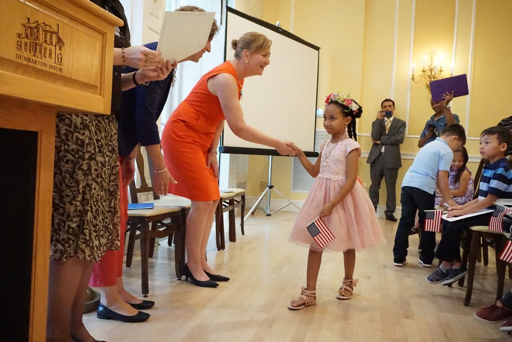 Young girl in a pink dress and flower crown carries a small American flag and shakes the hand of a Dame in an orange dress