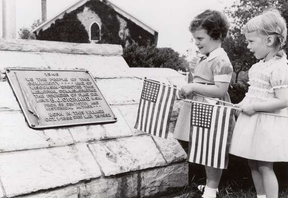 Black and white photo of two young children holding small US flags reading a plaque on a stone wall